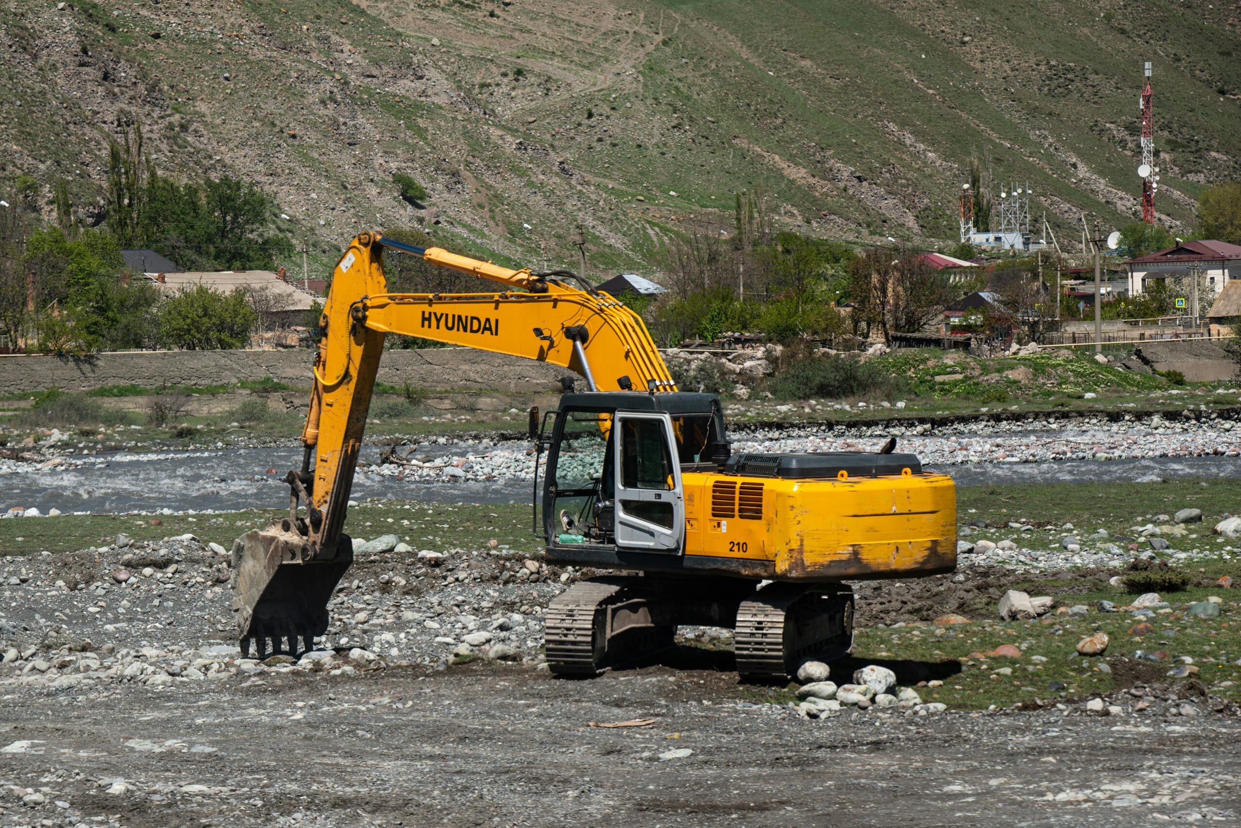 Yellow Hyundai excavator working on a rocky terrain near a river in a rural area. HD Hyundai Merger equipment